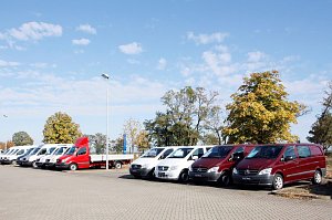 Reihe von Mercedes-Transportern und Vans, darunter ein roter Pritschenwagen, auf dem Autohaus-Parkplatz an einem sonnigen Tag mit blauem Himmel und Bäumen im Hintergrund.