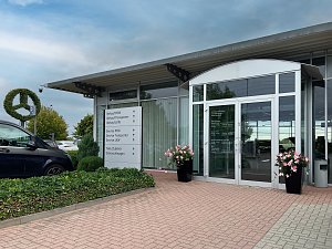 Eingangsbereich des Mercedes-Benz Autohauses in Bernburg mit gro&szlig;en Glasfronten. Davor ein wei&szlig;es Hinweisschild mit Richtungsangaben zu Verkauf, Service, Zubeh&ouml;r und Gebrauchtwagen, daneben Blumenk&uuml;bel.