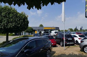 Blick &uuml;ber den Parkplatz eines Opel-Autohauses in S&ouml;mmerda. Im Vordergrund stehen viele verschiedene Fahrzeuge, darunter schwarze, rote und wei&szlig;e Modelle. Im Hintergrund ist das moderne Autohaus mit der markanten gelben Opel-Beschilderung zu sehen, umgeben von B&auml;umen und Flaggen. Der Himmel ist blau mit ein paar Wolken.