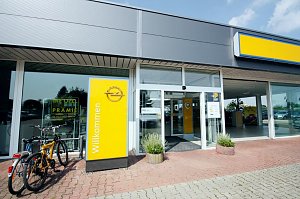 Eingang des Opel-Autohauses in S&ouml;mmerda. Links stehen zwei Fahrr&auml;der an einem Gel&auml;nder. Im Vordergrund ein gelbes Schild mit dem Opel-Logo und der Aufschrift Willkommen. Der Eingang ist verglast mit automatischen T&uuml;ren, daneben stehen Pflanzk&uuml;bel. Das Geb&auml;ude wirkt modern, mit gro&szlig;en Glasfronten und einem grauen Dach.
