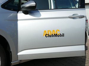 Nahaufnahme einer silbernen Autot&uuml;r mit der Beschriftung ADAC ClubMobil in gelber und schwarzer Schrift. Im Bild ist der linke Au&szlig;enspiegel, der T&uuml;rgriff und ein Teil des Hinterrads zu sehen. Das Fahrzeug steht auf einem gepflasterten Platz.