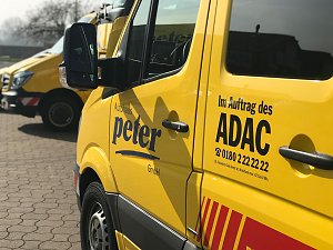 Gelber ADAC-Transporter vom Autohaus Peter, Nahaufnahme der Seitent&uuml;r mit den Logos Autohaus Peter und Im Auftrag des ADAC sowie Telefonnummer. Im Hintergrund weitere gelbe ADAC-Fahrzeuge, gepflasterter Hof und B&auml;ume.
