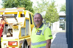 Matthias Köhler, Mitarbeiter des Autohaus Peter als ADAC Mobilitätspartner, steht in leuchtend gelber Dienstkleidung mit ADAC-Logo vor einem gelben ADAC-Pannenhilfefahrzeug. Im Hintergrund Bäume und ein moderner Gebäudeeingang.