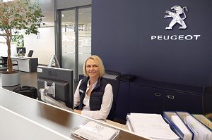 Freundliche Mitarbeiterin am Empfangsschalter eines Peugeot-Autohauses, im Hintergrund das Peugeot-Logo an der Wand, ein PC auf dem Schreibtisch und eine Pflanze links im Bild.