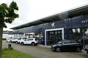 Peugeot Autohaus in G&ouml;ttingen mit gro&szlig;er Glasfassade und prominentem L&ouml;wen-Logo, mehrere wei&szlig;e und ein schwarzes Peugeot-Fahrzeug vor dem Geb&auml;ude geparkt.