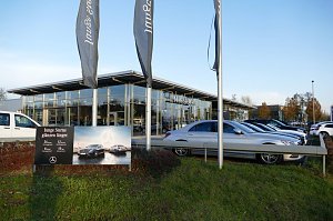 Mercedes-Benz Autohaus mit gro&szlig;er Glasfassade und davor geparkten Fahrzeugen, vor allem silberfarbene Limousinen. Im Vordergrund ein Schild mit der Aufschrift �Junge Sterne gl&auml;nzen l&auml;nger� und einem Bild von Mercedes-Fahrzeugen. Drei hohe Fahnenmasten mit Mercedes-Benz-Fahnen. Gr&uuml;nfl&auml;che mit Gras und Str&auml;uchern, blauer Himmel.