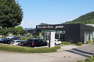 Au&szlig;enansicht des Mercedes-Benz Autohauses Peter in Sondershausen. Das moderne Geb&auml;ude mit schwarzer Fassade tr&auml;gt die Aufschrift 'Mercedes-Benz' und 'Peter'. Davor sind viele Autos auf dem Parkplatz geparkt, unter anderem ein schwarzer Van. Im Hintergrund sieht man gr&uuml;ne B&auml;ume und einen bewaldeten H&uuml;gel unter blauem Himmel.