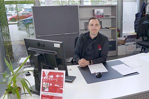 Mitarbeiter im Autohaus sitzt l&auml;chelnd an einem Schreibtisch mit Computer, Unterlagen und Pflanze, im Hintergrund Regale und Fenster mit Blick nach drau&szlig;en.