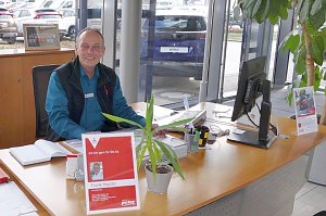 Ein Mitarbeiter sitzt freundlich l&auml;chelnd an einem Schreibtisch im Autohaus Peter in G&ouml;ttingen, mit Namensschild und Pflanzen auf dem Tisch, im Hintergrund ein Auto mit Peter-Nummernschild.