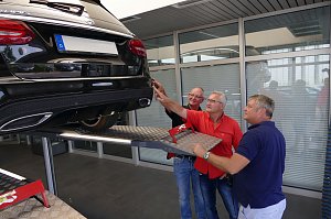 Drei M&auml;nner stehen in einem hellen Werkstattbereich unter dem Heck eines schwarzen Mercedes-AMG, der auf einer Hebeb&uuml;hne angehoben ist. Einer der M&auml;nner zeigt auf eine Stelle am Fahrzeug, die anderen beiden schauen interessiert zu.