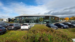 Mercedes-Benz Autohaus mit moderner Glasfassade und großem Mercedes-Logo, mehrere geparkte Fahrzeuge im Vordergrund, herbstliche Bäume und Wolkenhimmel im Hintergrund.