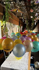 "Reihe bunter Luftballons mit der Aufschrift �Helau, Alaaf & Ahoi!� und dem Autohaus-Peter-Logo, befestigt an wei&szlig;en St&auml;ben, unter einer mit Pflanzen und Sternen dekorierten Decke.