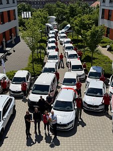 Fuhrparkaufstellung von oben: Reihe von DRK-Fahrzeugen, flankiert von Mitarbeitenden in roten Shirts. Im Vordergrund werden symbolisch Fahrzeugschl&uuml;ssel und Blumen &uuml;bergeben.