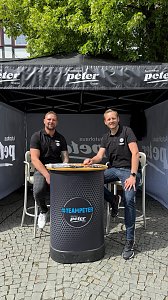 Infostand Autohaus Peter: Zwei Männer in schwarzen Polohemden mit Autohaus-Peter-Logo sitzen lächelnd an einem runden Stehtisch unter einem schwarzen Pavillon mit der Aufschrift Autohaus Peter Gruppe.