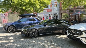 Premiumfahrzeuge in der Innenstadt: Ein blauer Mercedes-Benz SUV mit Dachbox und ein schwarzer Kombi stehen neben weiteren Fahrzeugen auf einem sonnigen Platz in einer Innenstadt mit Cafés und Geschäften.