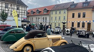 Mehrere Oldtimer stehen auf dem Marktplatz von Sondershausen vor einer Häuserzeile mit bunten Fassaden. Zu sehen sind historische Fahrzeuge in verschiedenen Farben und Karosserieformen unter anderem ein cremefarbenes Cabriolet im Vordergrund. Menschen schlendern zwischen den Autos.