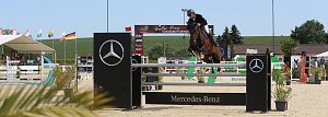 Das Bild zeigt einen Springreiter in Immenrode, der mit seinem Pferd &uuml;ber ein Hindernis mit Mercedes-Benz-Werbung springt. Im Hintergrund sieht man eine Reitturnier-Arena mit verschiedenen Hindernissen, Fahnen mehrerer L&auml;nder und Zuschauerbereichen. Das Wetter ist sonnig, und es gibt einige Pflanzen und Dekorationen um die Arena.