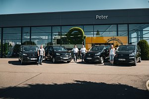 Vor einem modernen Autohaus mit dem Namen Peter stehen vier schwarze Opel-Transporter in einer Reihe. Vier Personen drei Männer und eine Frau posieren jeweils neben den Fahrzeugen. An der Glasfassade ist das Opel-Logo und ein gelber Hintergrund zu sehen.