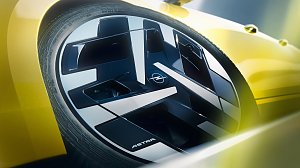 Detailaufnahme einer Fahrzeugfelge mit einem geometrisch gestalteten, schwarz-silbernen Felgendesign. In der Mitte befindet sich das Opel-Logo. Am unteren Rand des Felgenblatts steht ASTRA. Das gelbe Fahrzeugheck ist im Hintergrund unscharf zu erkennen, akzentuiert durch weiche Lichteff
