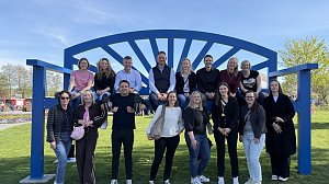 Gruppenfoto: Gro&szlig;e blaue XXL-Bank, darauf sitzen mehrere Personen, einige stehen davor und l&auml;cheln in die Kamera.