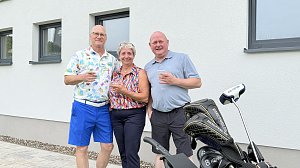Drei G&auml;ste stehen mit Getr&auml;nken neben einem Golftrolley vor dem Clubhaus.