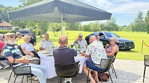 G&auml;ste sitzen an einem Tisch unter Sonnenschirmen mit Blick auf den Golfplatz und ausgestellte Fahrzeuge.
