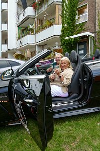Zwei Frauen, die l&auml;chelnd in einem schwarzen Mercedes-Cabriolet mit ge&ouml;ffnetem Verdeck sitzen und mit Getr&auml;nken ansto&szlig;en.