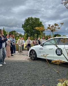 Eine gr&ouml;&szlig;ere Gruppe von Frauen, die sich im Freien unter bew&ouml;lktem Himmel versammelt haben und aufmerksam einen wei&szlig;en Mercedes-Benz CLA betrachten.