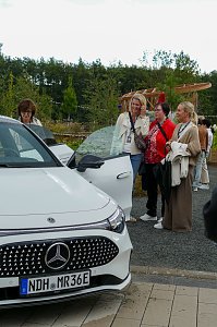 Eine Gruppe von Frauen, die sich im Freien unter bew&ouml;lktem Himmel versammelt haben und aufmerksam einen wei&szlig;en Mercedes-Benz CLA betrachten.