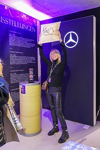 Eine Frau in dunkler Kleidung, die freudig ein Kissen mit der Aufschrift She�s Mercedes vor einer Mercedes-Eventwand in die H&ouml;he h&auml;lt.