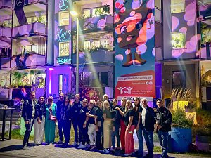 Mitarbeiter des Autohauses, die im Freien vor einer farbenfroh illuminierten Geb&auml;udefassade mit gro&szlig;fl&auml;chiger Pop-Art-Projektion f&uuml;r ein Gruppenfoto posieren.