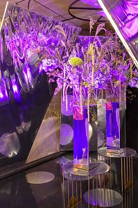 Hohe Glasvasen mit floralen Arrangements, die auf kleinen runden Tischen unter intensivem, lilafarbenem Event-Licht vor spiegelnden W&auml;nden stehen.