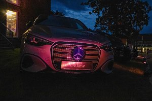 Die Frontansicht eines Mercedes-Benz, der in der D&auml;mmerung im Freien steht und dessen K&uuml;hlergrill sowie das Markenlogo lila beleuchtet sind.