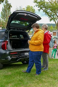 Zwei Frauen in farbigen Jacken, die im Freien auf einer Wiese stehen und den ge&ouml;ffneten Kofferraum eines schwarzen Mercedes-Fahrzeugs betrachten.