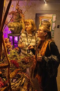Zwei Frauen, die in einem stimmungsvoll beleuchteten Innenraum aufmerksam ein kunstvolles, florales Arrangement betrachten.