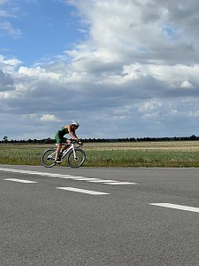Radstrecke beim Triathlon. Ein Athlet im gr&uuml;nen Einteiler und mit wei&szlig;em Helm f&auml;hrt auf einem hellen Rennrad vorbei. Dahinter erstreckt sich eine weite Feldlandschaft unter einem leicht bew&ouml;lkten Himmel.