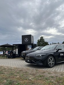 Zwei schwarze Mercedes-Benz Fahrzeuge stehen vor dem Promotion-Stand. Ein Stehtisch mit Sonnenschirm l&auml;dt Besucher zum Verweilen und Informieren ein.