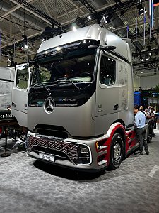 Silberner Mercedes-Benz Actros Lkw von vorne links in einer Messehalle. Die untere Seitenverkleidung und die Radl&auml;ufe sind dunkelrot lackiert. Ein Mann in blauem Hemd steht rechts neben dem Fahrzeug. Der Lkw steht auf grauem Teppichboden.