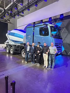 F&uuml;nf Personen stehen l&auml;chelnd nebeneinander vor einem hellblauen Mercedes-Benz Betonmischer auf einer Messeb&uuml;hne. Die gro&szlig;e Mischtrommel hinten ist wei&szlig;-blau gestreift.