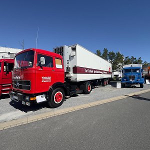 Roter historischer Mercedes-Benz Sattelzug von vorne links auf einem asphaltierten Platz. Der wei&szlig;e Auflieger hat einen braunen Streifen. Rote Felgen. Im Hintergrund rechts steht ein blauer Oldtimer-Lkw unter wolkenlosem, blauem Himmel.