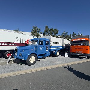 Blauer Oldtimer-Lkw mit langer Motorhaube und beigen Felgen von vorne links. Er zieht einen wei&szlig;-blauen Anh&auml;nger mit MICHELIN-Aufschrift. Rechts daneben steht ein orangefarbener Lkw.