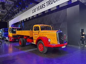 Gelber Mercedes-Benz Hauben-Lkw mit roten Kotfl&uuml;geln und rotem Fahrgestell schr&auml;g von vorne auf einer Messeb&uuml;hne. Er hat eine offene, gelbe Ladefl&auml;che. Im Hintergrund stehen moderne Lkws unter einem Banner mit 130 YEARS TRUCKS.