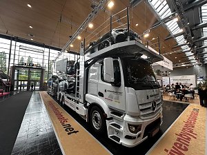 Wei&szlig;er Mercedes-Benz Autotransporter schr&auml;g von vorne in einer gro&szlig;en Messehalle. Mehrere Pkws sind auf den Rampen &uuml;ber dem Fahrerhaus und auf dem Anh&auml;nger geladen. Der Lkw steht auf einer beigen Ausstellungsfl&auml;che mit roter Schrift.