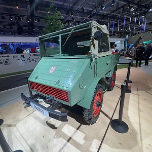 Gr&uuml;ner historischer Mercedes-Benz Unimog schr&auml;g von vorne auf einem hellen Holzboden auf einer Messe. Er hat auff&auml;llige, rote Felgen, eine eckige Front und ein beiges Stoffverdeck. Im Hintergrund stehen Messebesucher. Schwarze Absperrstangen davor.