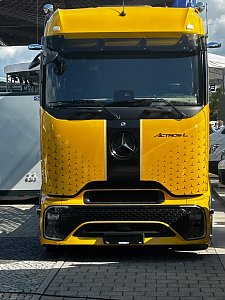 Gelber Mercedes-Benz Lkw direkt von vorne fotografiert. In der Mitte der Front verl&auml;uft ein breiter, schwarzer L&auml;ngsstreifen mit dem Markenlogo. Das Fahrzeug steht auf einem gepflasterten Au&szlig;enbereich.