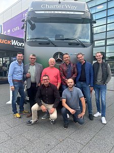 Acht M&auml;nner posieren l&auml;chelnd vor einem gro&szlig;en, grauen Mercedes-Benz Lkw auf einem Au&szlig;enbereich. Sechs M&auml;nner stehen aufrecht nebeneinander, w&auml;hrend zwei M&auml;nner davor in der Hocke sitzen. Oben auf dem Lkw steht der wei&szlig;e Schriftzug CharterWay.