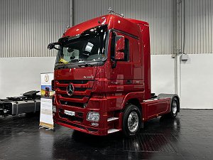 Dunkelroter Mercedes-Benz Actros Lkw schr&auml;g von vorne in einer Halle. Das fahrbereite Fahrerhaus gl&auml;nzt im Scheinwerferlicht. Hinten ist das freie Fahrgestell ohne Auflieger zu sehen.