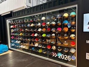 Gro&szlig;e Vitrine in einem schwarzen Container voller bunter Baseballkappen. Viele verschiedene Caps sind ordentlich in Reihen nebeneinander aufgereiht. Unten rechts auf der Glasscheibe steht die wei&szlig;e Jahreszahl 2026 mit Pfeilen.