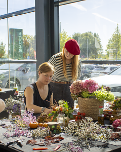 Zwei Frauen gestalten an einem Tisch herbstliche Blumenarrangements aus Hortensien, Zweigen, Beeren und kleinen K&uuml;rbissen.