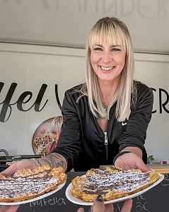 L&auml;chelnde Mitarbeiterin eines Gastronomiestands reicht zwei frisch gebackene Waffeln mit Schokolade und Puderzucker &uuml;ber die Theke.